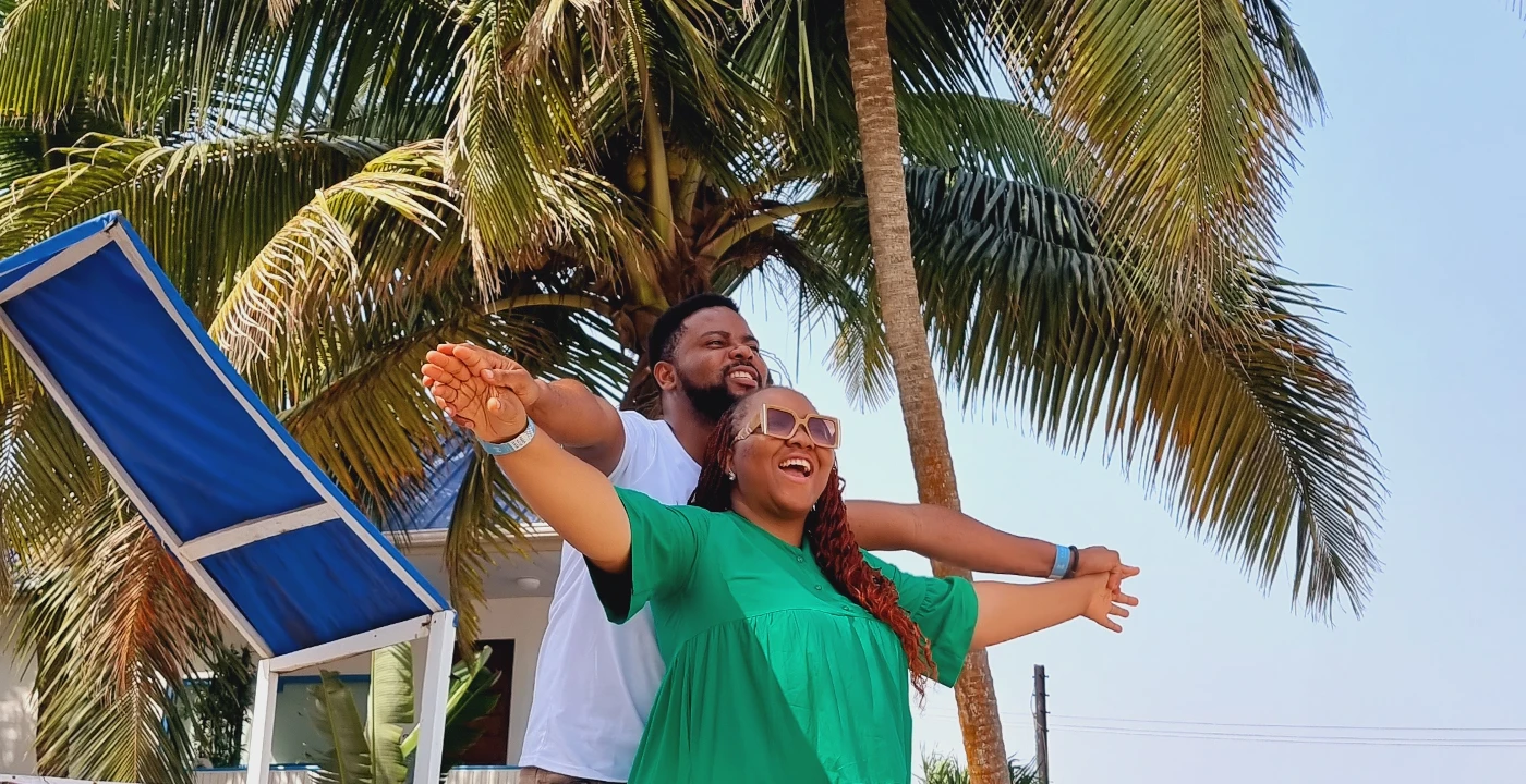 A couple recreating a Titanic pose on a wooden deck surrounded by palm trees, enjoying their romantic honeymoon in Nigeria.