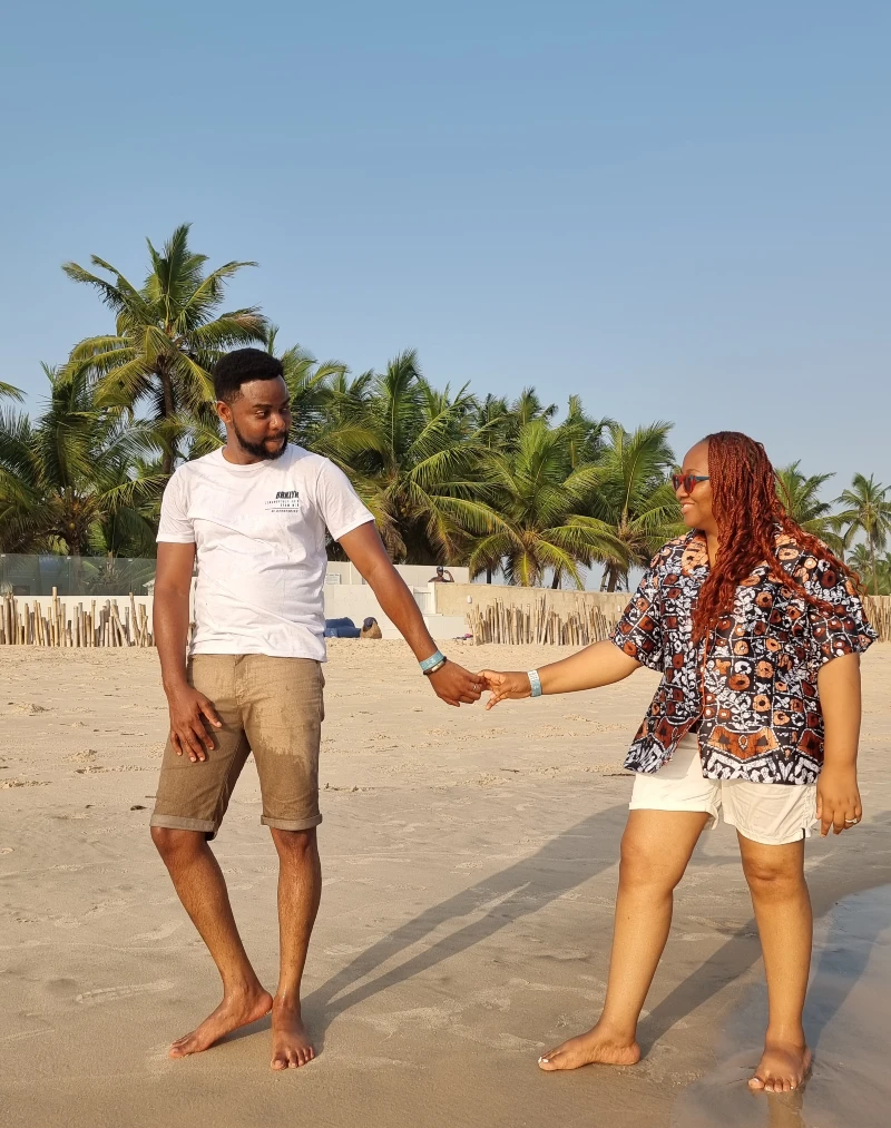 A couple holding hands during a romantic walk on a sandy beach in Nigeria, with palm trees in the background.