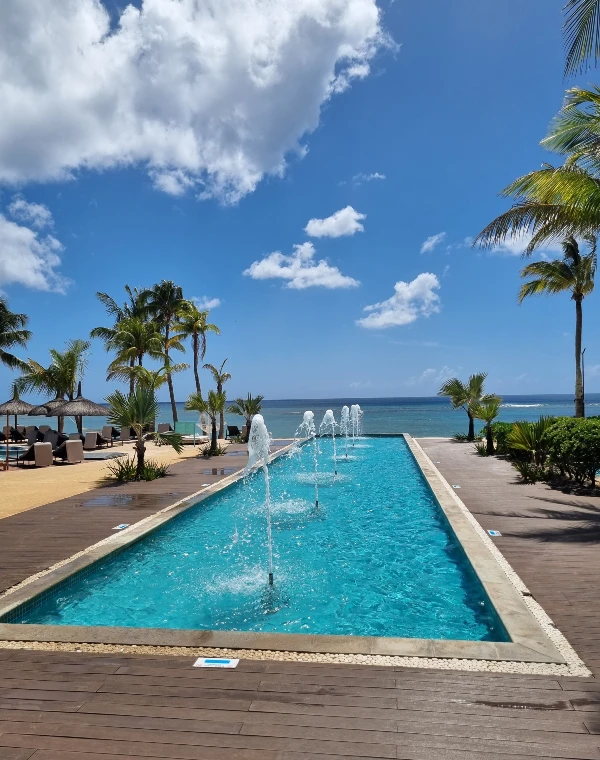 A luxurious poolside view at a tropical honeymoon destination, with palm trees, water fountains, and a clear blue sky overlooking the ocean.