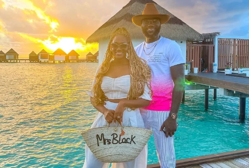 A smiling couple posing at sunset by the turquoise waters of a luxurious overwater bungalow, holding a customized bag that says 'Mrs. Black.