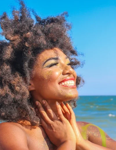 A woman with natural curly hair smiling under the bright sun on a beach with the ocean in the background.