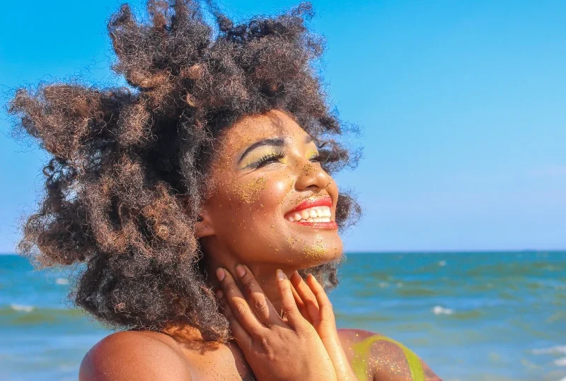 A woman with natural curly hair smiling under the bright sun on a beach with the ocean in the background.