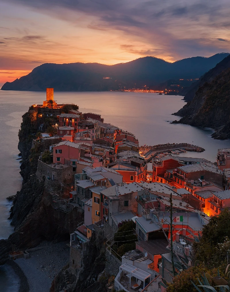 A scenic sunset view over a cliffside village with colorful houses overlooking the sea, surrounded by mountains.