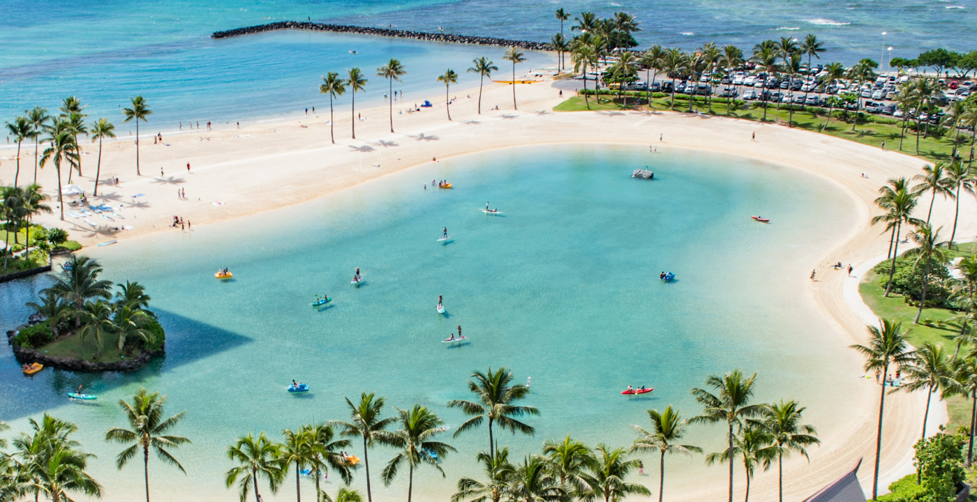 A calm tropical beach lagoon surrounded by palm trees, with people kayaking and paddleboarding on the clear turquoise waters.