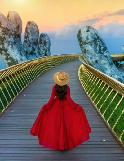 A woman in a red dress walking along the Golden Bridge in Da Nang, Vietnam, with giant stone hands supporting the bridge, set against a sunset sky.