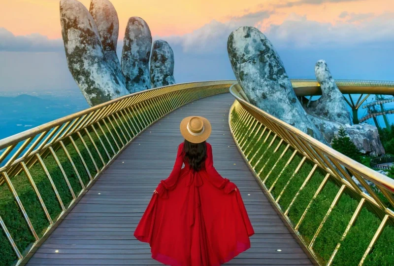 A woman in a red dress walking along the Golden Bridge in Da Nang, Vietnam, with giant stone hands supporting the bridge, set against a sunset sky.