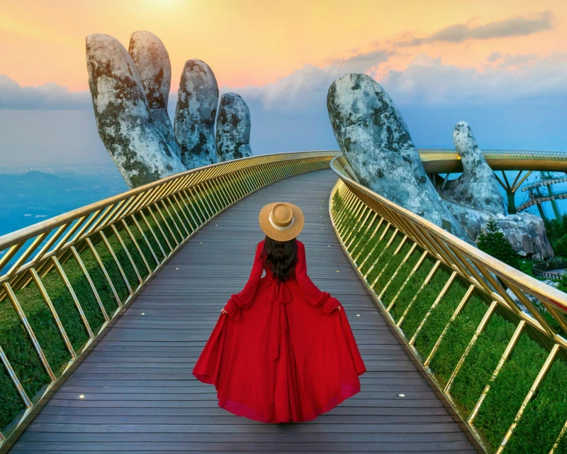 A woman in a red dress walking along the Golden Bridge in Da Nang, Vietnam, with giant stone hands supporting the bridge, set against a sunset sky.