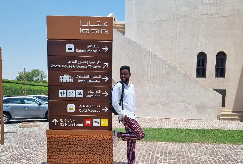 Man standing next to a signpost in Katara Cultural Village, Doha, highlighting tourist attractions with Lery Hago Travels