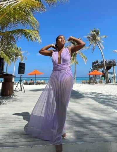 A woman in a flowing lavender dress enjoying the sunshine at a tropical all-inclusive beach resort, surrounded by palm trees and beach umbrellas.