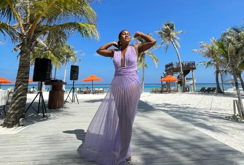 A woman in a flowing lavender dress enjoying the sunshine at a tropical all-inclusive beach resort, surrounded by palm trees and beach umbrellas.