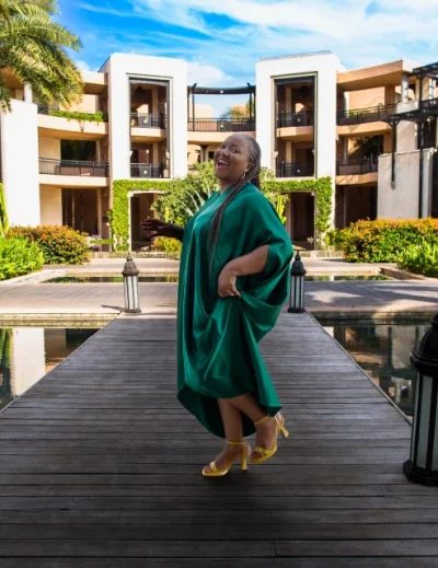 Woman in a green dress standing on a boardwalk at a luxury resort, surrounded by lush greenery and elegant buildings.