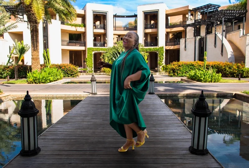 Woman in a green dress standing on a boardwalk at a luxury resort, surrounded by lush greenery and elegant buildings.