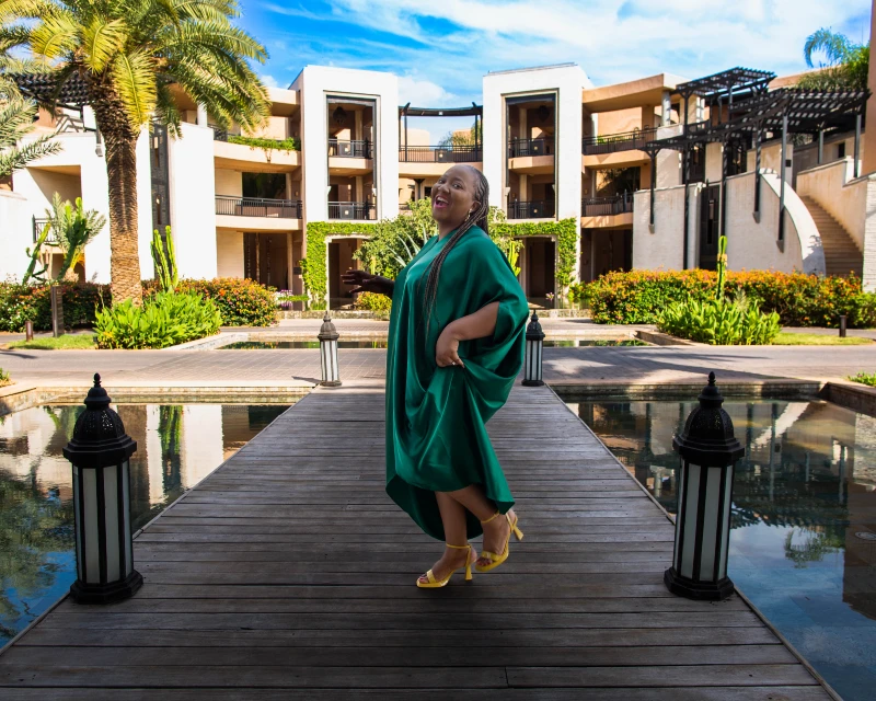 Woman in a green dress standing on a boardwalk at a luxury resort, surrounded by lush greenery and elegant buildings.