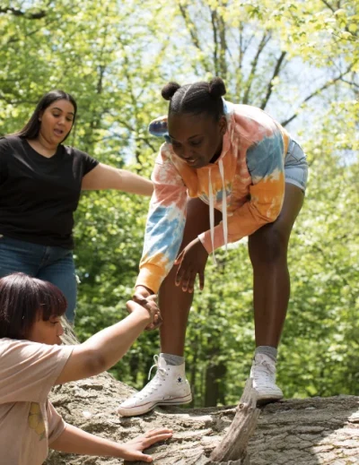 "A group of people helping each other climb over a log in a forest during a family vacation, showcasing teamwork and adventure. Your essential family vacation packing list: what to bring when travelling with kids of all ages - Lery Hago Travels."