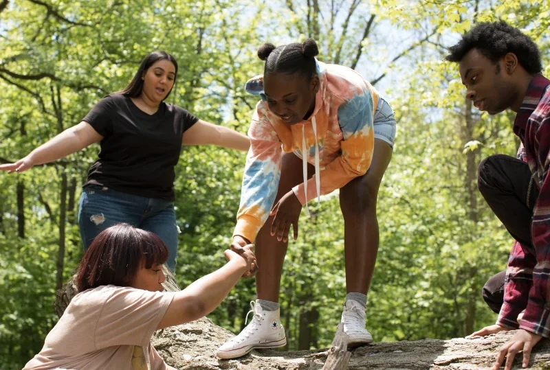 "A group of people helping each other climb over a log in a forest during a family vacation, showcasing teamwork and adventure. Your essential family vacation packing list: what to bring when travelling with kids of all ages - Lery Hago Travels."