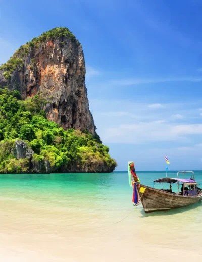 "Traditional long-tail boat on a serene beach with a towering limestone cliff in Thailand, perfect for a tropical getaway."