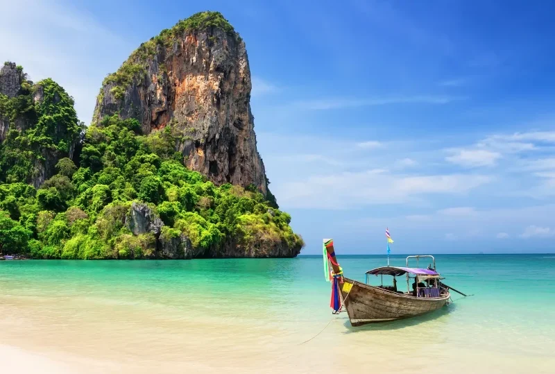 "Traditional long-tail boat on a serene beach with a towering limestone cliff in Thailand, perfect for a tropical getaway."