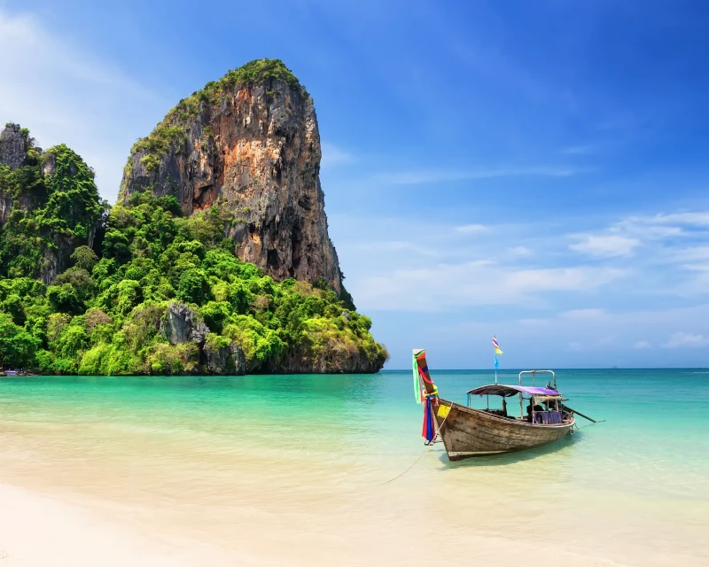 "Traditional long-tail boat on a serene beach with a towering limestone cliff in Thailand, perfect for a tropical getaway."