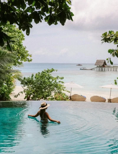 "A woman in a hat relaxing in an infinity pool with a view of a tropical beach and calm ocean waters."