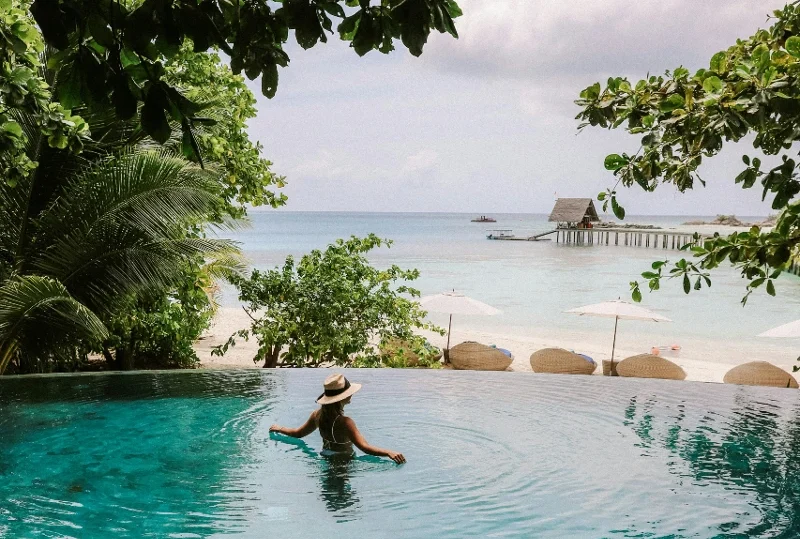 "A woman in a hat relaxing in an infinity pool with a view of a tropical beach and calm ocean waters."