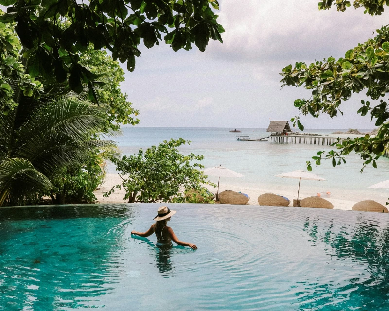 "A woman in a hat relaxing in an infinity pool with a view of a tropical beach and calm ocean waters."