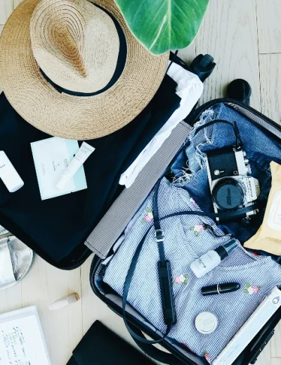 Open suitcase with must-have solo travel essentials, including a sun hat, camera, skincare items, sandals, and a notepad for a stress-free journey with Lery Hago Travels.