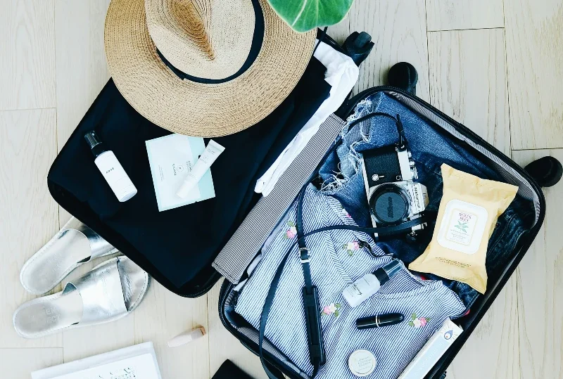 Open suitcase with must-have solo travel essentials, including a sun hat, camera, skincare items, sandals, and a notepad for a stress-free journey with Lery Hago Travels.