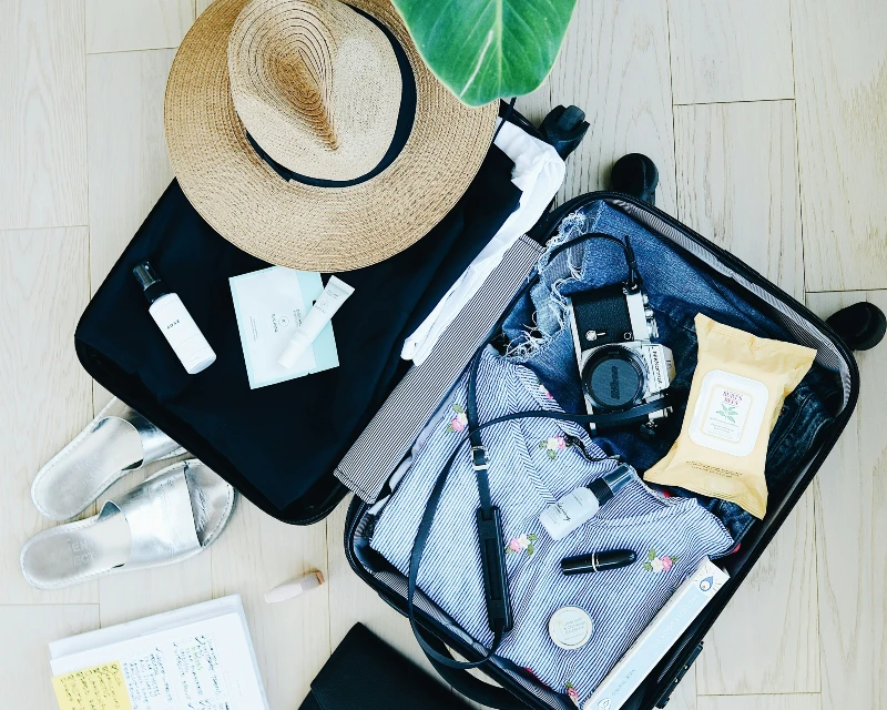 Open suitcase with must-have solo travel essentials, including a sun hat, camera, skincare items, sandals, and a notepad for a stress-free journey with Lery Hago Travels.