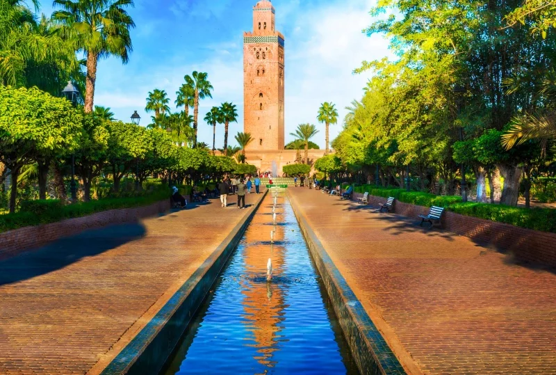 Scenic view of the Koutoubia Mosque in Marrakech, Morocco, with a tranquil water feature and lush gardens, captured on a sunny day.