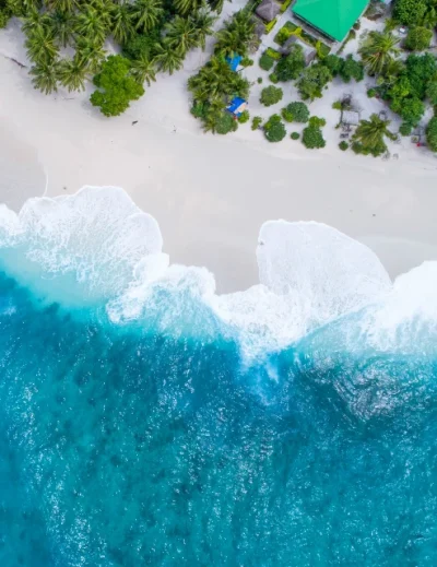 "A mesmerizing aerial view of waves crashing onto a Maldives beach, bordered by lush greenery and a secluded resort, perfect for first-timers with Lery Hago Travels."