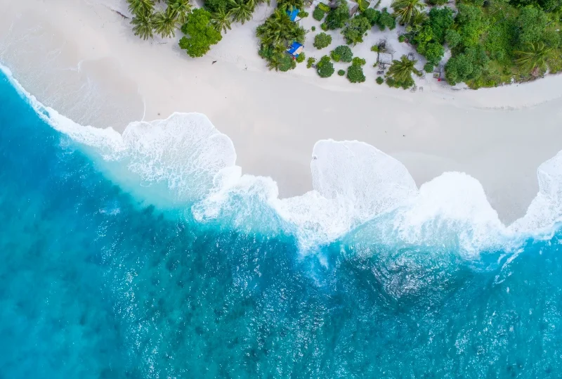 "A mesmerizing aerial view of waves crashing onto a Maldives beach, bordered by lush greenery and a secluded resort, perfect for first-timers with Lery Hago Travels."