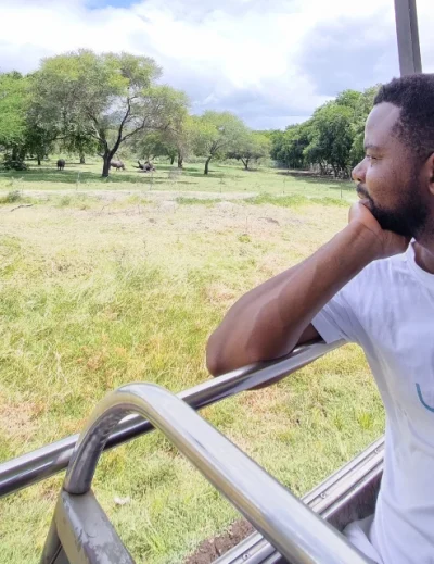 "Man wearing a Lery Hago Travels t-shirt enjoying a safari ride, looking out at lush green trees and wildlife under a bright blue sky."