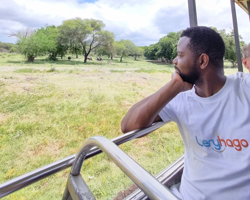 "Man wearing a Lery Hago Travels t-shirt enjoying a safari ride, looking out at lush green trees and wildlife under a bright blue sky."