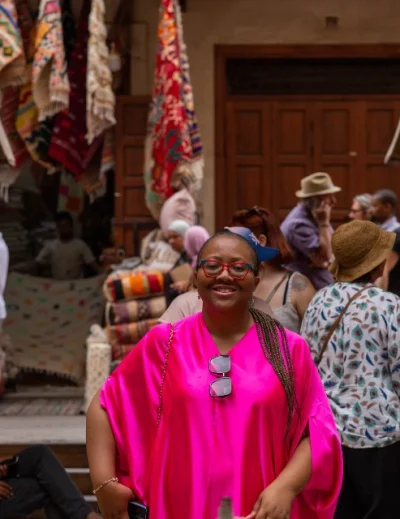 "A smiling woman in a bright pink outfit exploring a colorful market in Egypt, surrounded by tourists and traditional rugs on display."