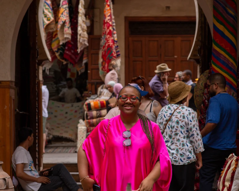 "A smiling woman in a bright pink outfit exploring a colorful market in Egypt, surrounded by tourists and traditional rugs on display."