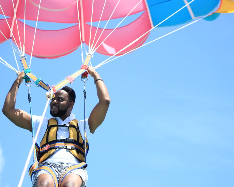 "A man enjoying parasailing under a vibrant parachute against a clear blue sky, showcasing an adventurous holiday experience with Lery Hago Travels."