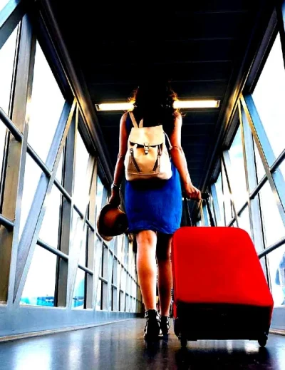 A woman in a blue dress with a backpack and red suitcase walking through an airport boarding gate for international travel.