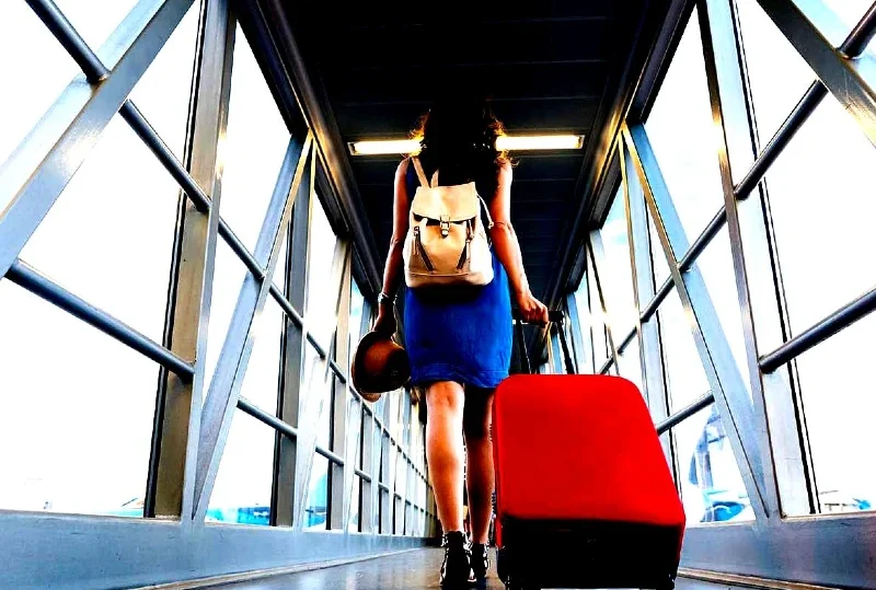 A woman in a blue dress with a backpack and red suitcase walking through an airport boarding gate for international travel.