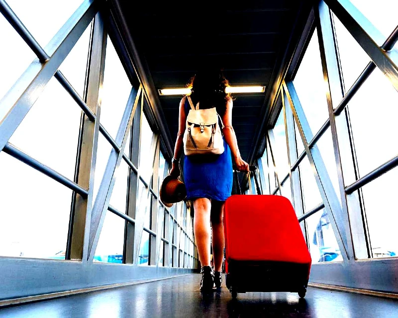 A woman in a blue dress with a backpack and red suitcase walking through an airport boarding gate for international travel.