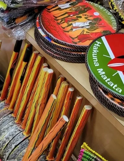 A collection of traditional Kenyan souvenirs, including hand-painted drums, decorative plates, and wooden carvings, displayed in a vibrant market stall.