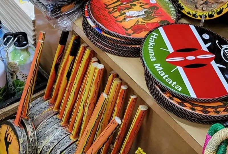 A collection of traditional Kenyan souvenirs, including hand-painted drums, decorative plates, and wooden carvings, displayed in a vibrant market stall.