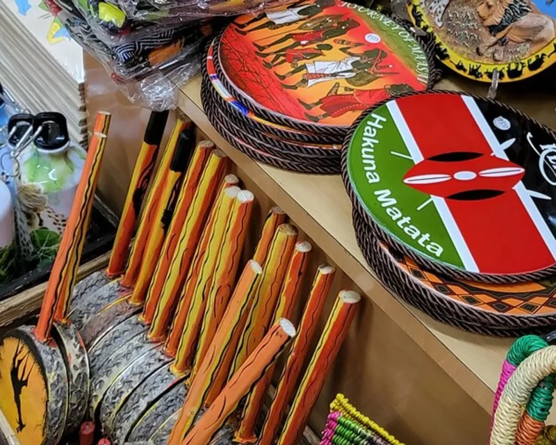 A collection of traditional Kenyan souvenirs, including hand-painted drums, decorative plates, and wooden carvings, displayed in a vibrant market stall.