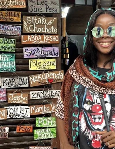 "A smiling woman in a patterned scarf standing in front of colorful Swahili phrase boards in Zanzibar, Tanzania."