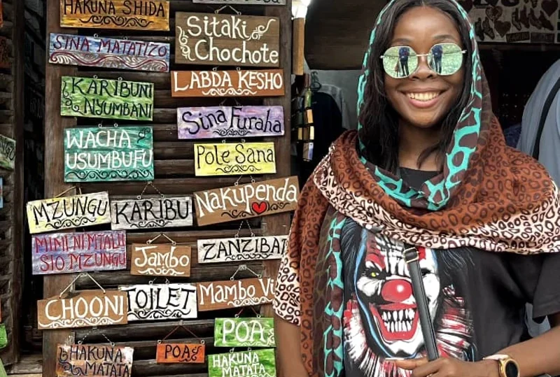 "A smiling woman in a patterned scarf standing in front of colorful Swahili phrase boards in Zanzibar, Tanzania."