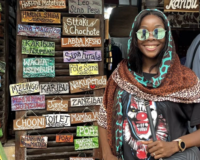 "A smiling woman in a patterned scarf standing in front of colorful Swahili phrase boards in Zanzibar, Tanzania."