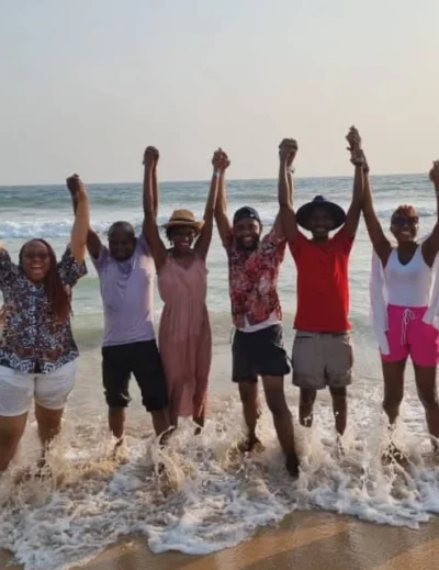 "A cheerful group of friends holding hands while enjoying the waves on a sunny beach in Lagos, capturing the essence of Detty December fun with Lery Hago Travels."