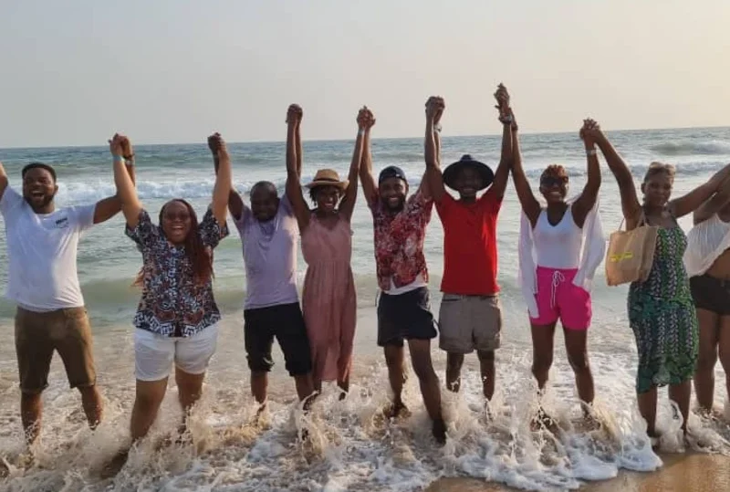 "A cheerful group of friends holding hands while enjoying the waves on a sunny beach in Lagos, capturing the essence of Detty December fun with Lery Hago Travels."