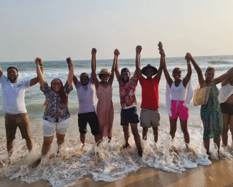 "A cheerful group of friends holding hands while enjoying the waves on a sunny beach in Lagos, capturing the essence of Detty December fun with Lery Hago Travels."