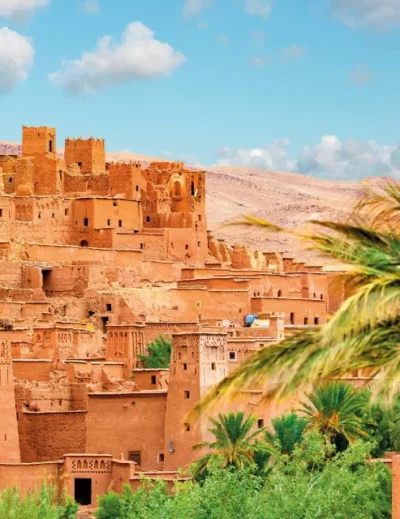 "The ancient clay city of Ait Benhaddou in Morocco, surrounded by lush palm trees under a bright blue sky."