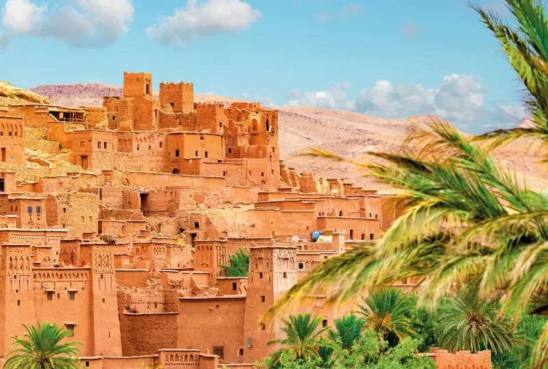 "The ancient clay city of Ait Benhaddou in Morocco, surrounded by lush palm trees under a bright blue sky."